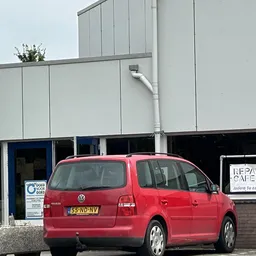 Voorkant van kringloopcentrum met witte gevel, rode VW auto geparkeerd ervoor en 'Repair Cafe' bord zichtbaar