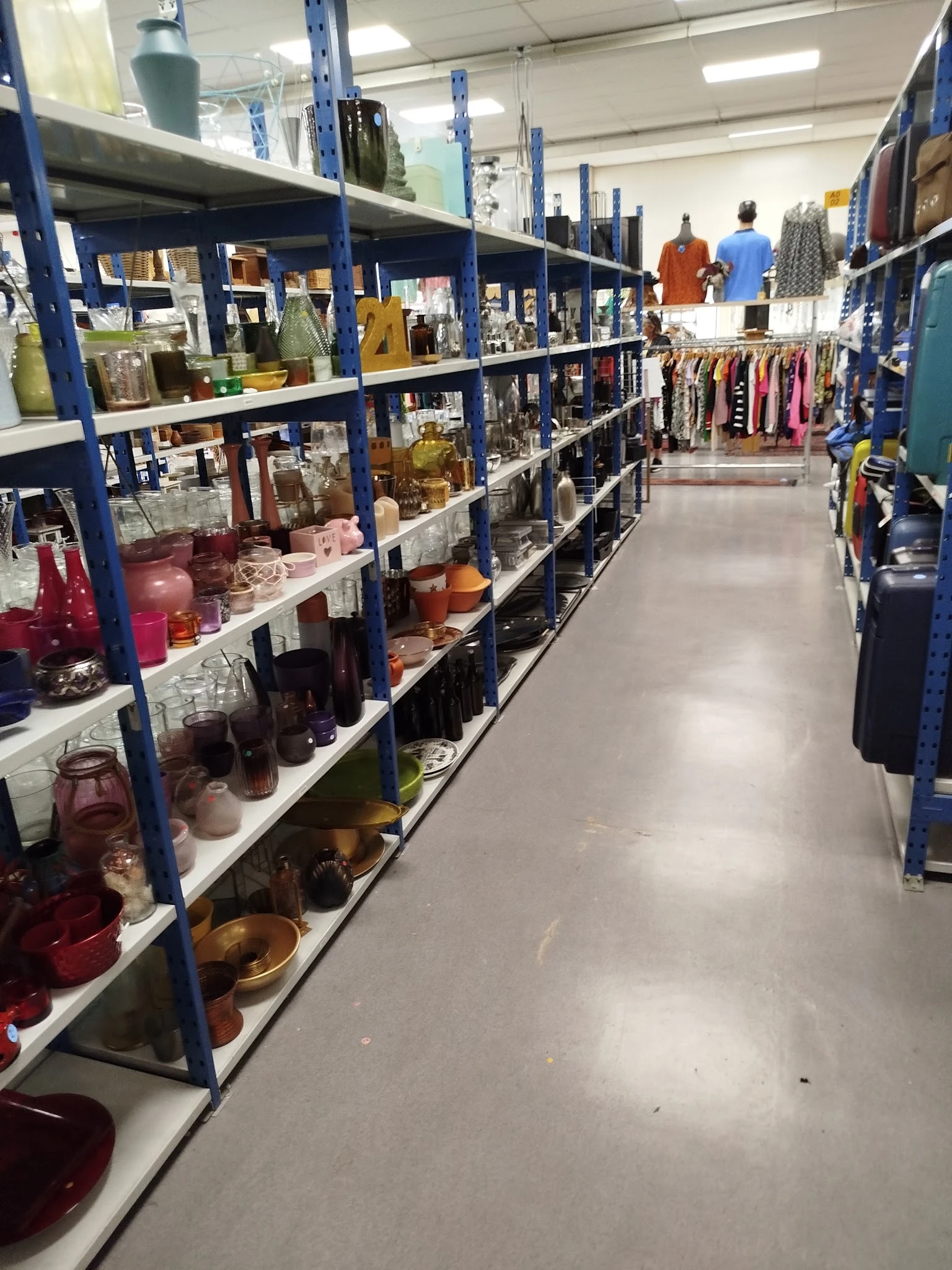 Gangpad tussen blauwe stellingen met huishoudelijke spullen en kleding in kringloopwinkel