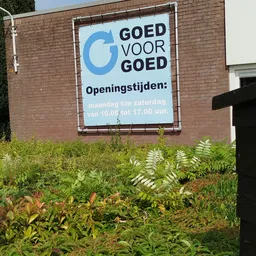 Openingsbord 'Goed voor Goed' met openingstijden op een bakstenen gevel met begroeiing