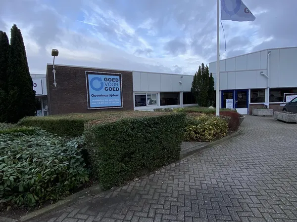 Gevel van kringloopcentrum met blauwe logo en ingang, omgeven door struiken en plaveisel