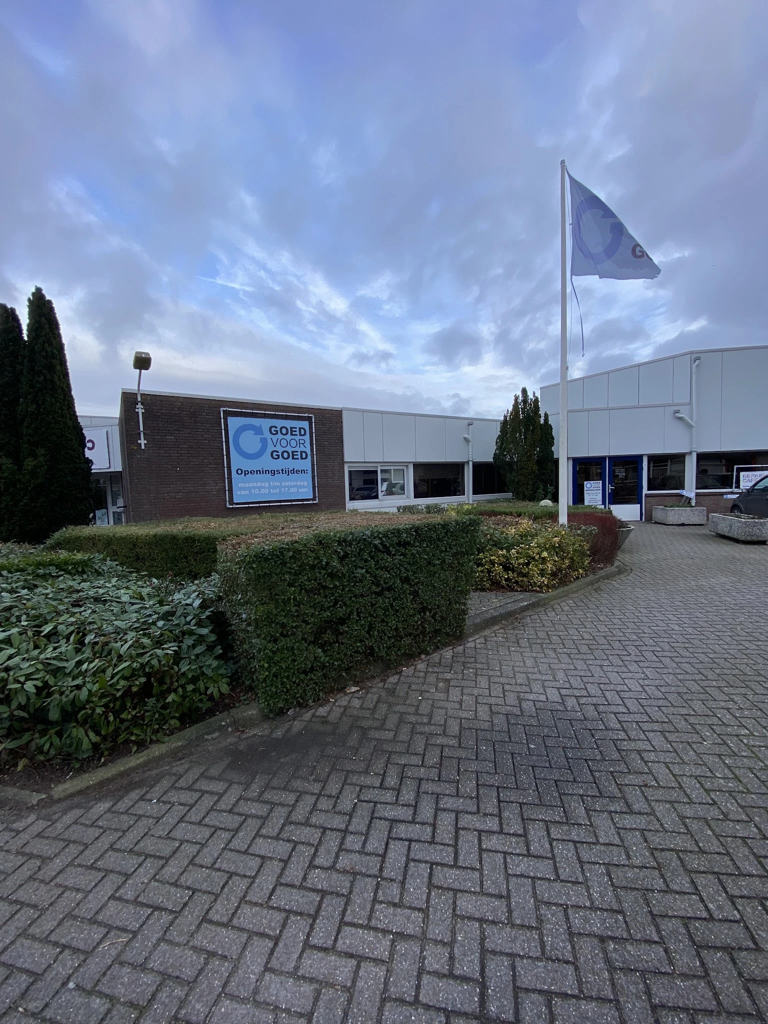 Gevel van kringloopcentrum met blauwe logo en ingang, omgeven door struiken en plaveisel
