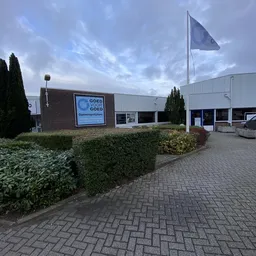 Gevel van kringloopcentrum met blauwe logo en ingang, omgeven door struiken en plaveisel