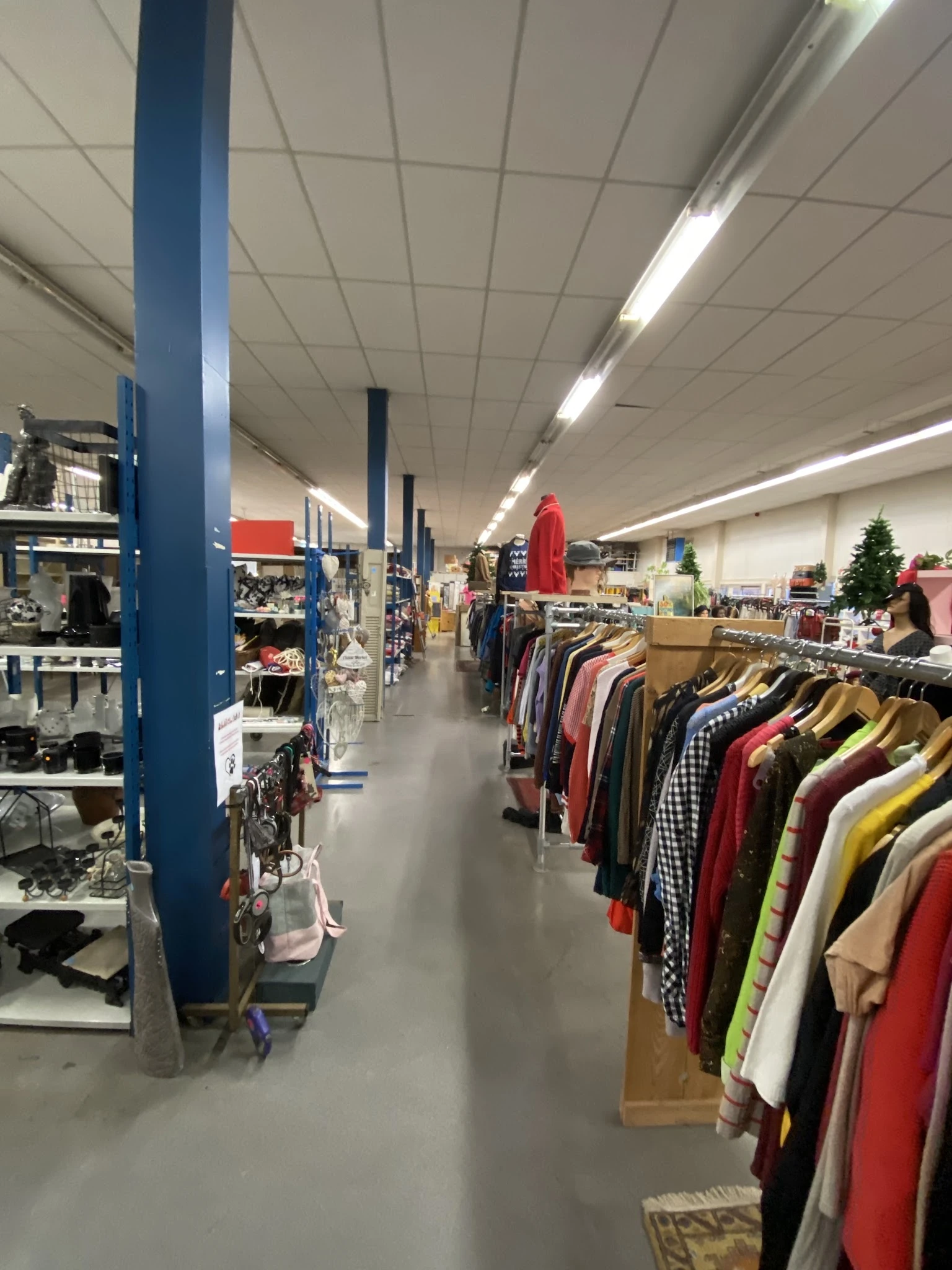 Binnenaanzicht van kringloopwinkel met rekken tweedehands kleding, blauwe steunpilaren en verlichte plafondpanelen