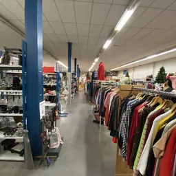 Binnenaanzicht van kringloopwinkel met rekken tweedehands kleding, blauwe steunpilaren en verlichte plafondpanelen