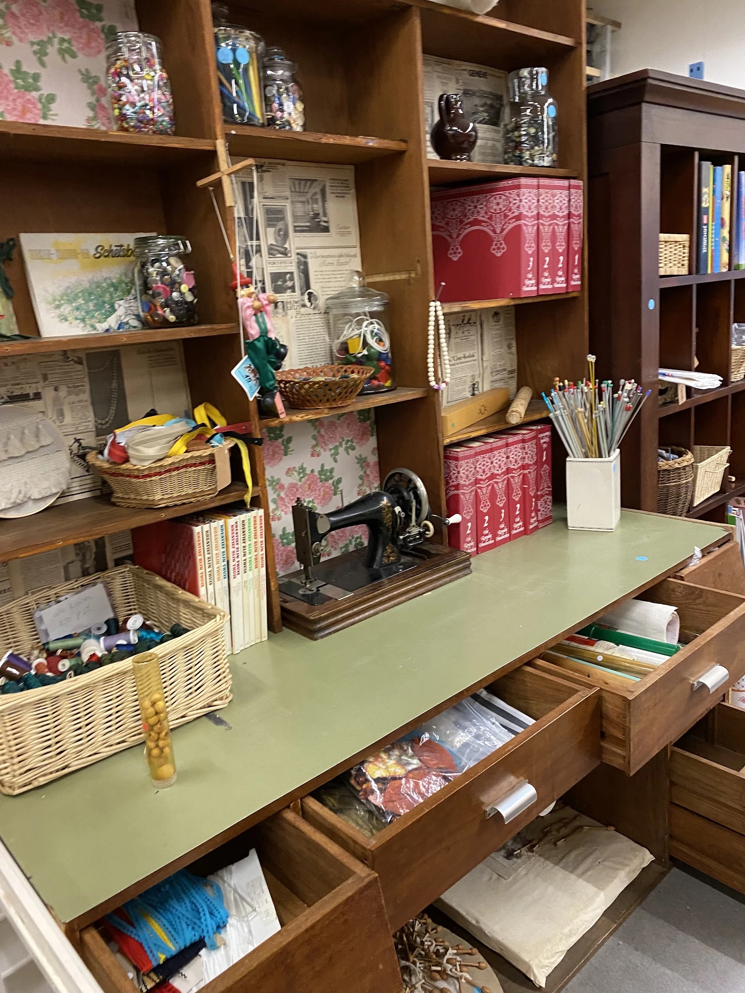 Winkelinterieur met houten stellingen vol spullen, bureauwerk en decoratie; op de tafel staat een oude naaimachine