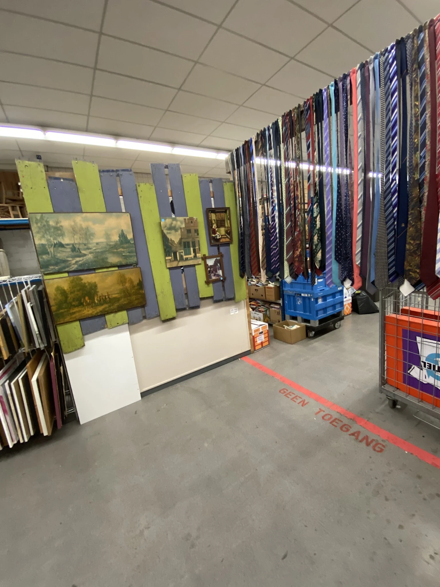 Winkelvloer met stellingen vol kunstwerk, lijsten en gordijnen in kringloopwinkel