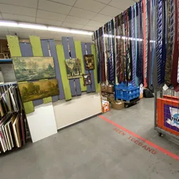 Winkelvloer met stellingen vol kunstwerk, lijsten en gordijnen in kringloopwinkel