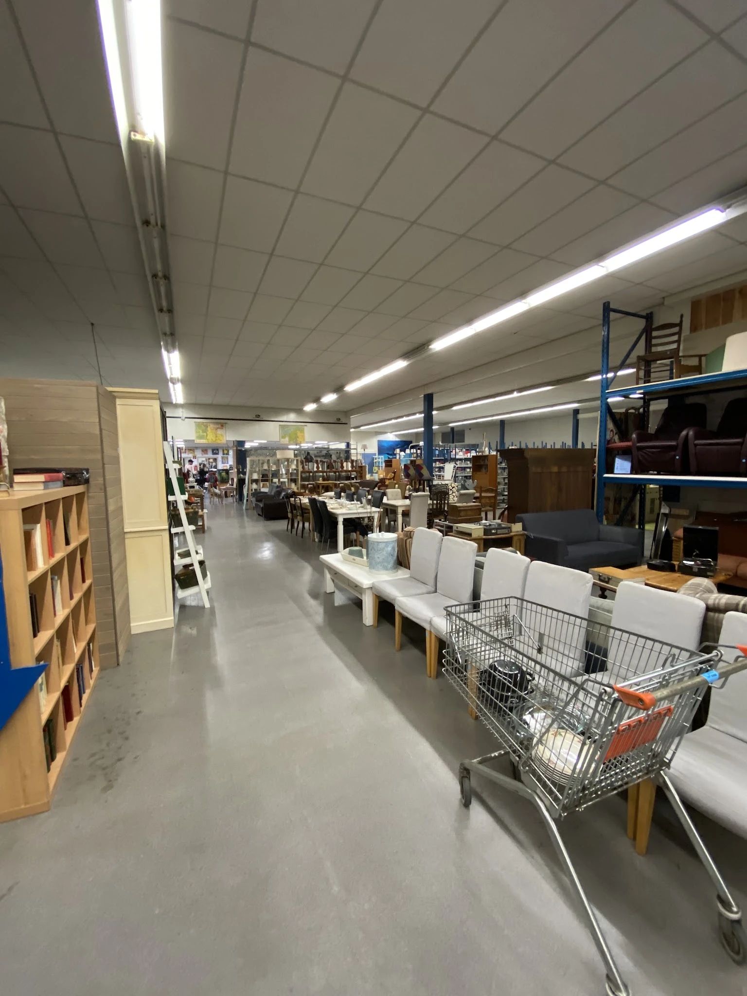 Binnenaanzicht van kringloopwinkel met stellingen, tafels met witte stoelen en winkelwagen op de vloer