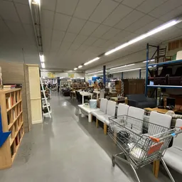 Binnenaanzicht van kringloopwinkel met stellingen, tafels met witte stoelen en winkelwagen op de vloer