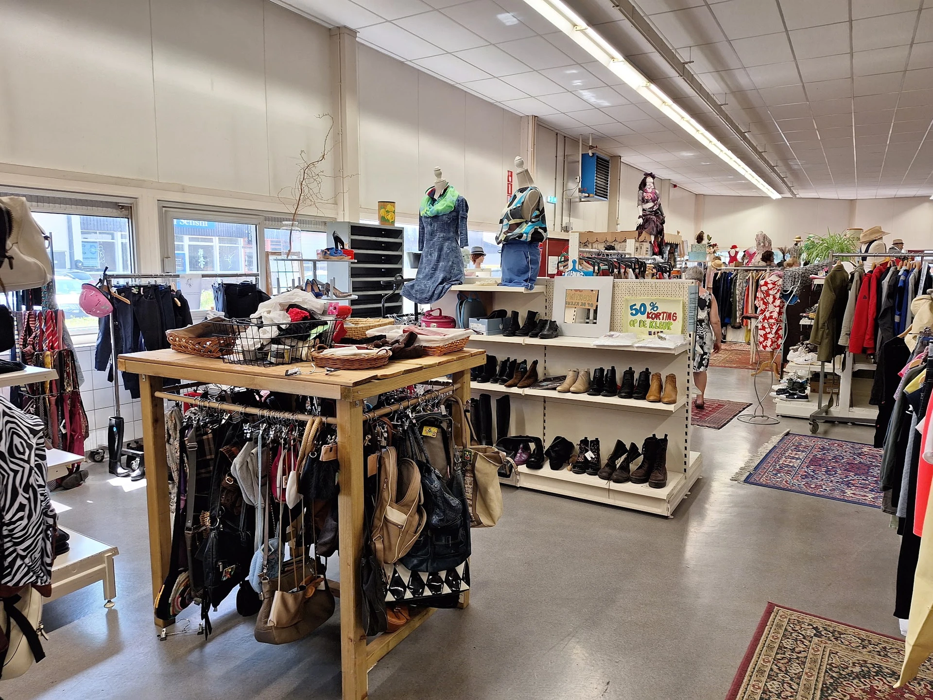 Overzicht van winkelruimte met kledingrekken, tafels met kleding, schoenen en accessoires, mannequins, en natuurlijk licht va