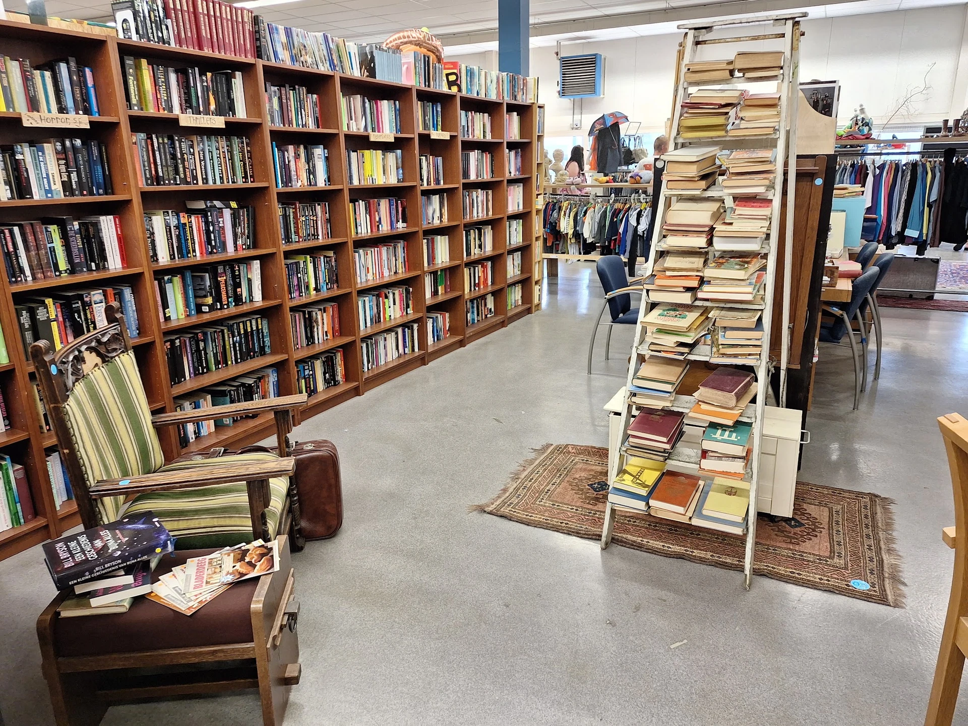 Winkelvloer met houten boekenkasten vol boeken, een leeszetel, en een bewegwijzering naar het kledinggedeelte
