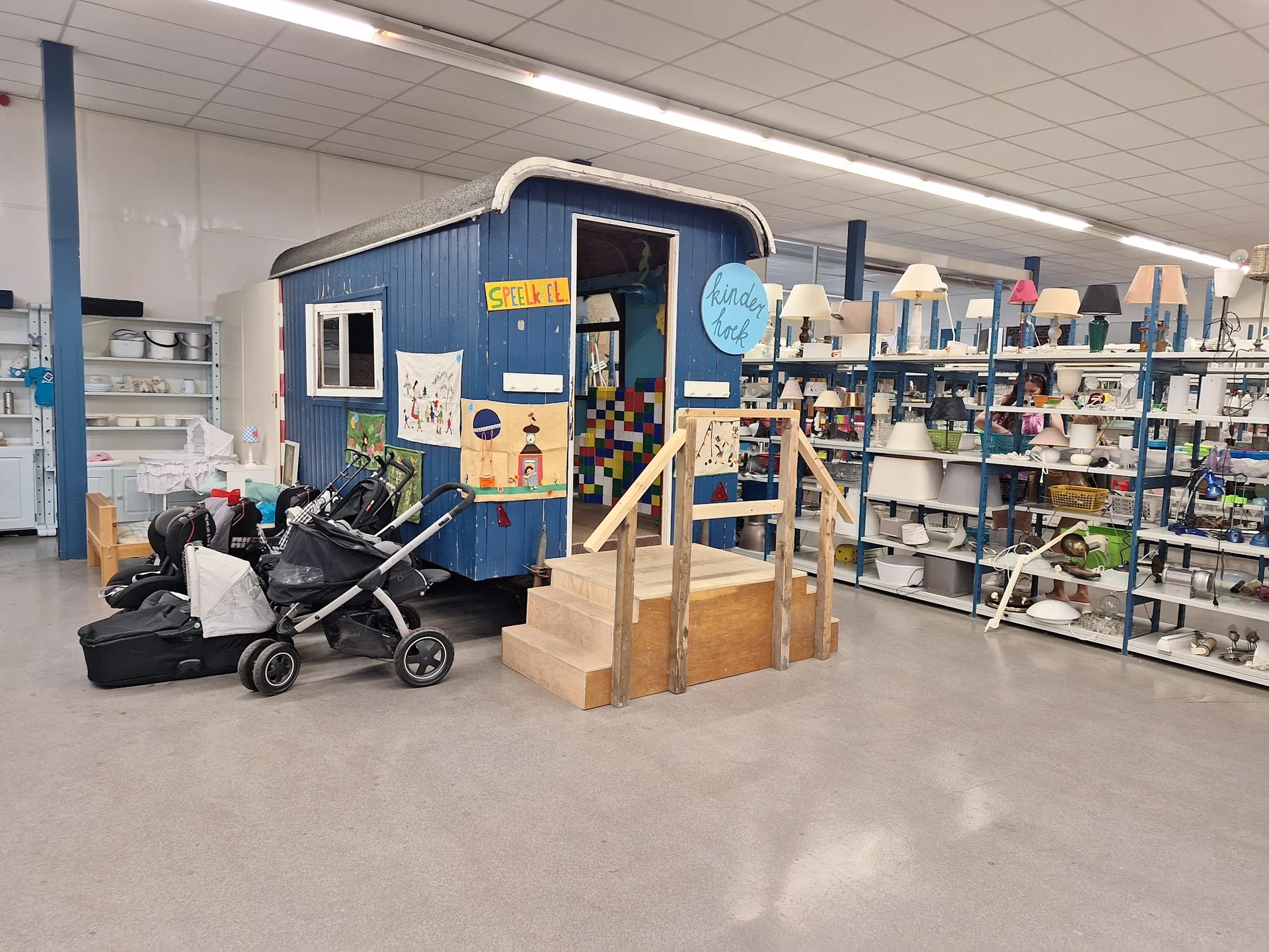 Winkelvloer met blauwe aanhangwagen, kinderwagen, houten speeltoestel en rekken met spullen