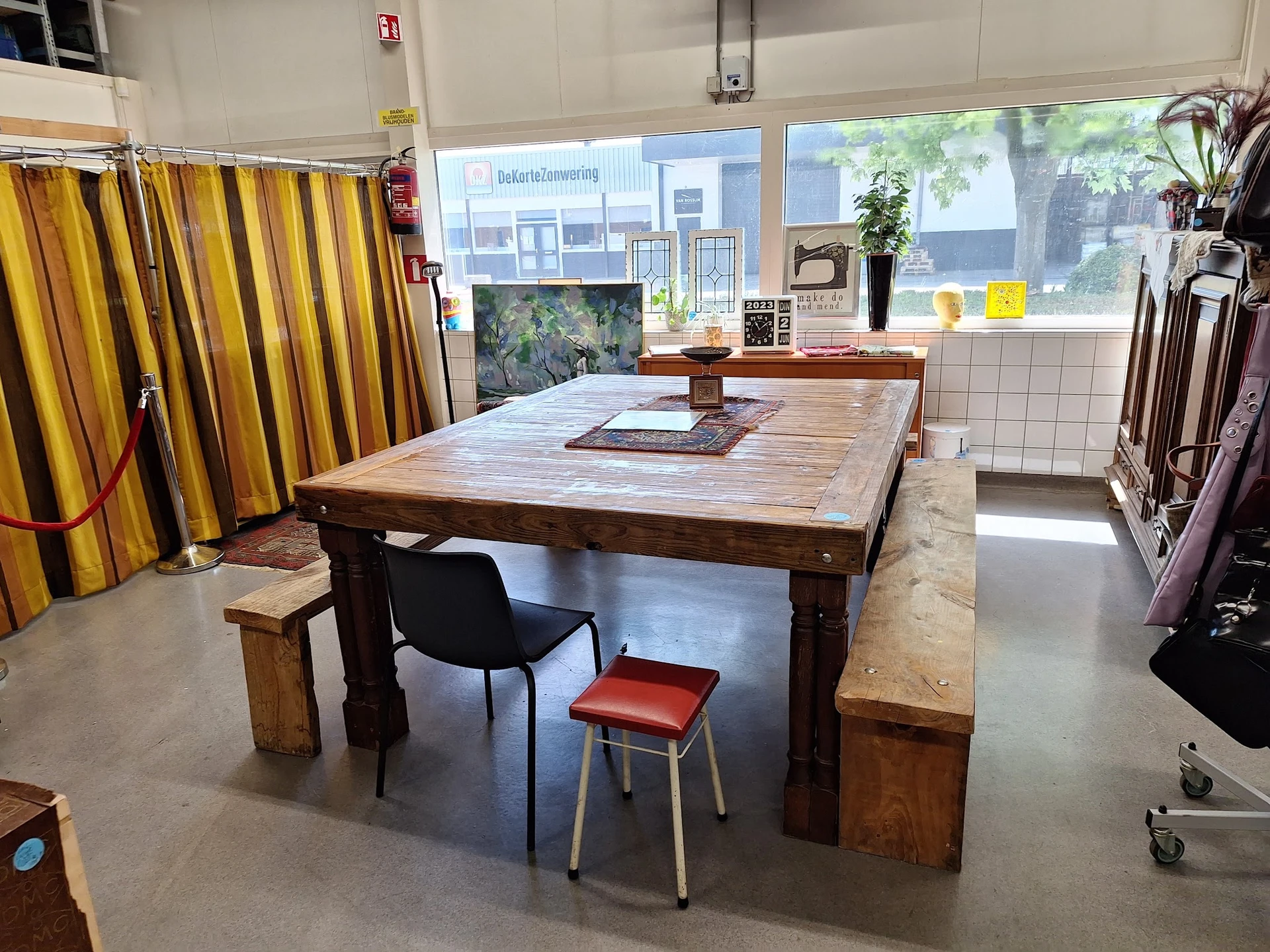 Werkruimte in kringloopwinkel met grote houten tafel, stoelen en geel gestreepte gordijn aan linkerkant
