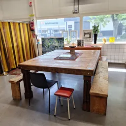 Werkruimte in kringloopwinkel met grote houten tafel, stoelen en geel gestreepte gordijn aan linkerkant