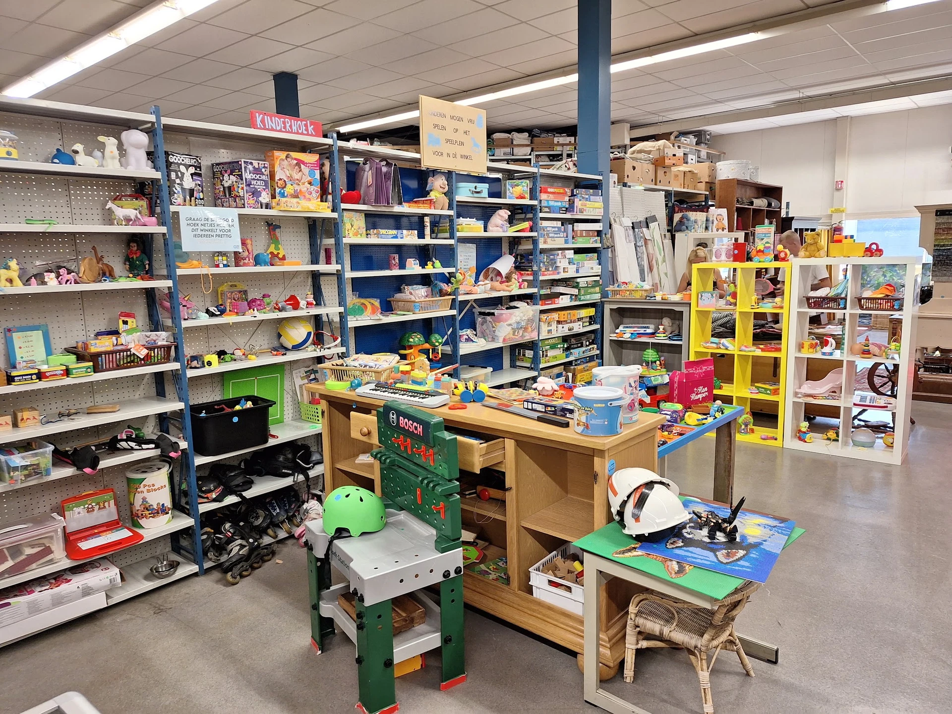 Winkelvloer met stellingen vol speelgoed, boeken en huishoudartikelen; in het midden een werktafel met groene werkbank