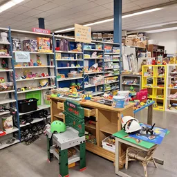 Winkelvloer met stellingen vol speelgoed, boeken en huishoudartikelen; in het midden een werktafel met groene werkbank