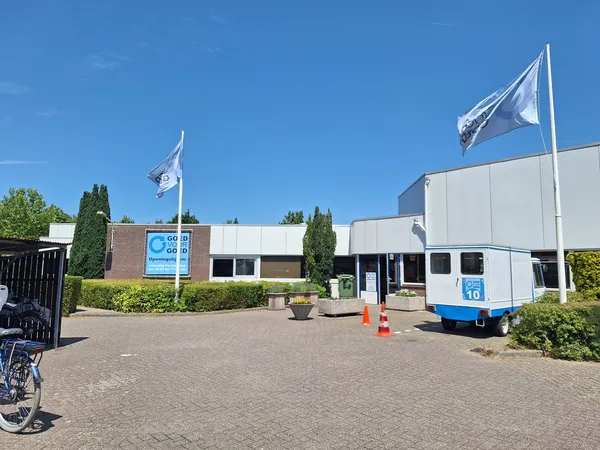 Exterieur van kringloopwinkel met bakstenen gevel, witte zijvleugel, blauwe vlaggen en groene hagen