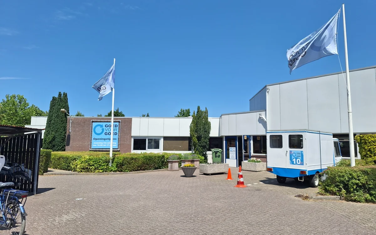 Exterieur van kringloopwinkel met bakstenen gevel, witte zijvleugel, blauwe vlaggen en groene hagen