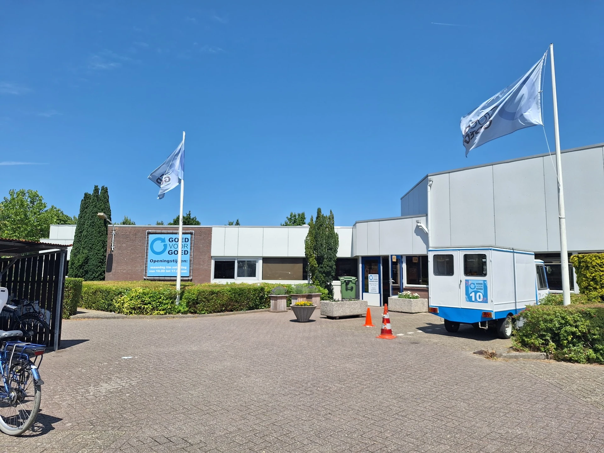 Exterieur van kringloopwinkel met bakstenen gevel, witte zijvleugel, blauwe vlaggen en groene hagen