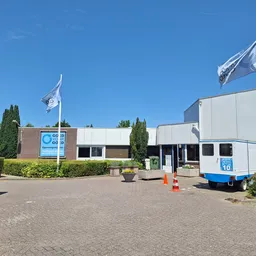 Exterieur van kringloopwinkel met bakstenen gevel, witte zijvleugel, blauwe vlaggen en groene hagen