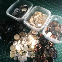 Plastic bakjes met diverse losse knopen in verschillende kleuren en materialen, uitgestald op groene ondergrond