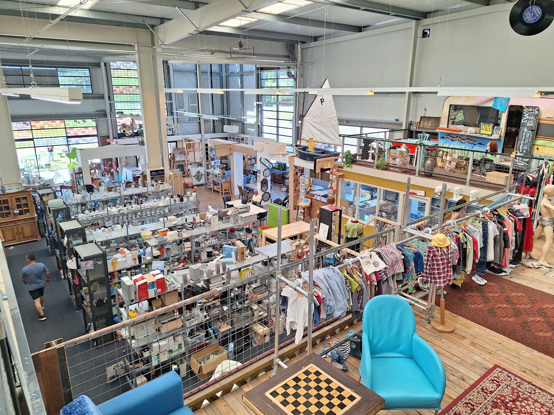 Overzicht van kringloopwinkel met stellingen vol spullen, kledingrekken en zithoeken met blauwe stoelen