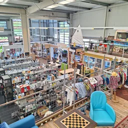 Overzicht van kringloopwinkel met stellingen vol spullen, kledingrekken en zithoeken met blauwe stoelen
