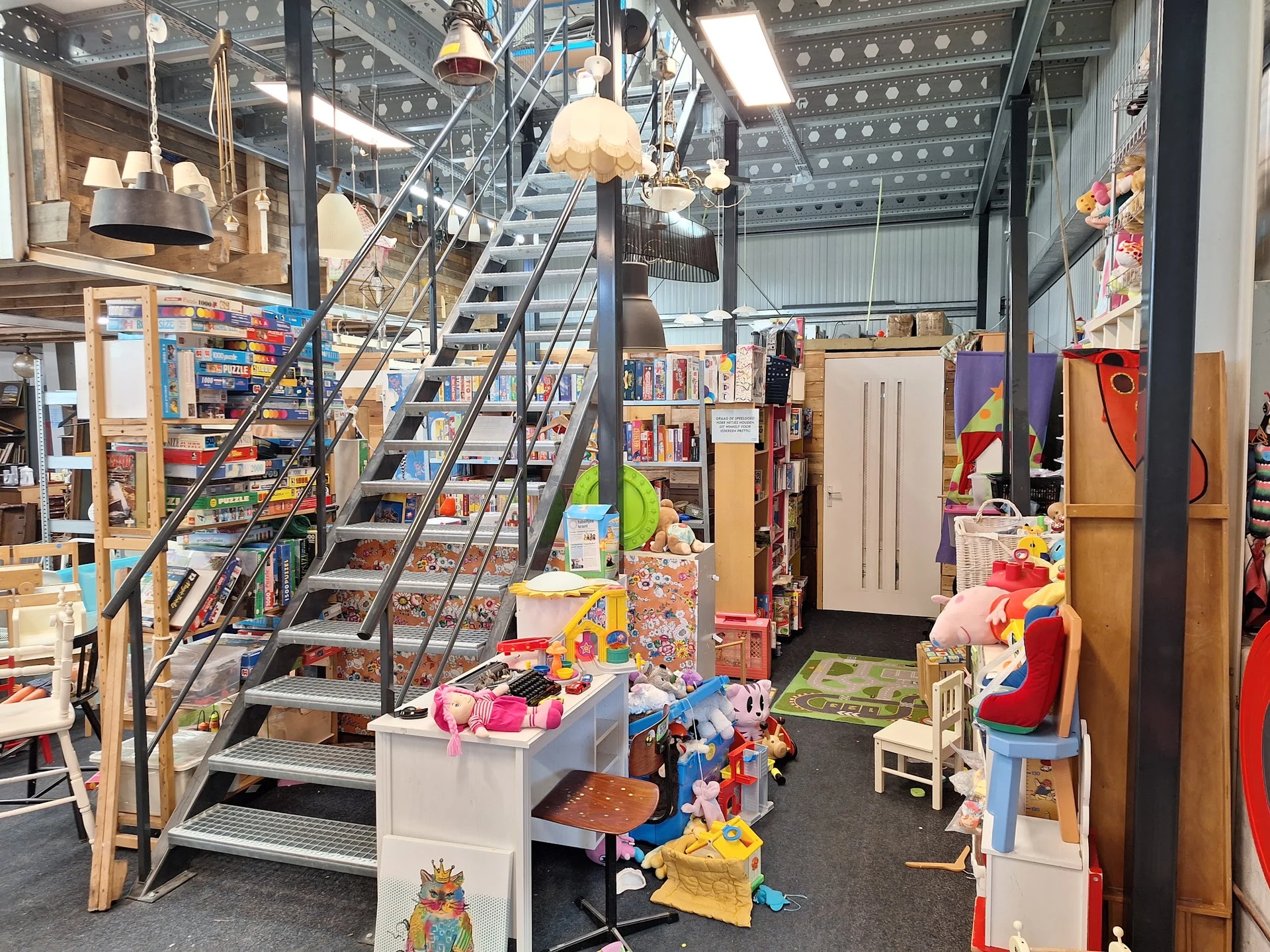 Winkelvloer van kringloopcentrum met trap, stellingen met speelgoed, boeken en diverse spullen, hanglampen en deuren zichtbaa