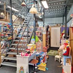 Winkelvloer van kringloopcentrum met trap, stellingen met speelgoed, boeken en diverse spullen, hanglampen en deuren zichtbaa