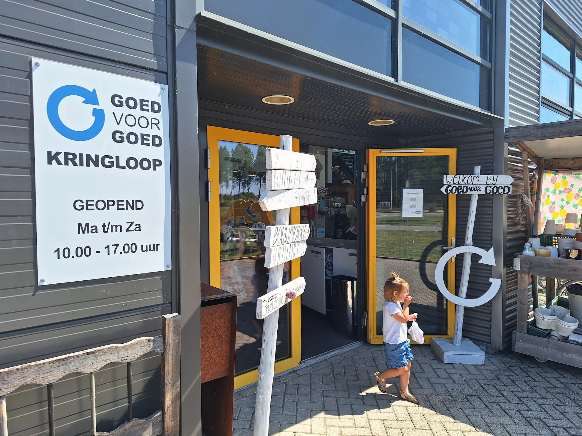 Voorgevel van kringloopwinkel met geel-zwarte deur, winkelnaam en openingstijden op bord, kind op stoep
