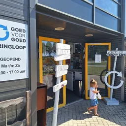 Voorgevel van kringloopwinkel met geel-zwarte deur, winkelnaam en openingstijden op bord, kind op stoep