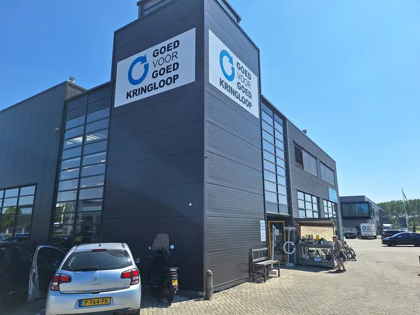 Zwart kringloopcentrum met glazen ramen en blauw logo 'Goed voor Goed Kringloop' op de voorgevel, auto geparkeerd ervoor