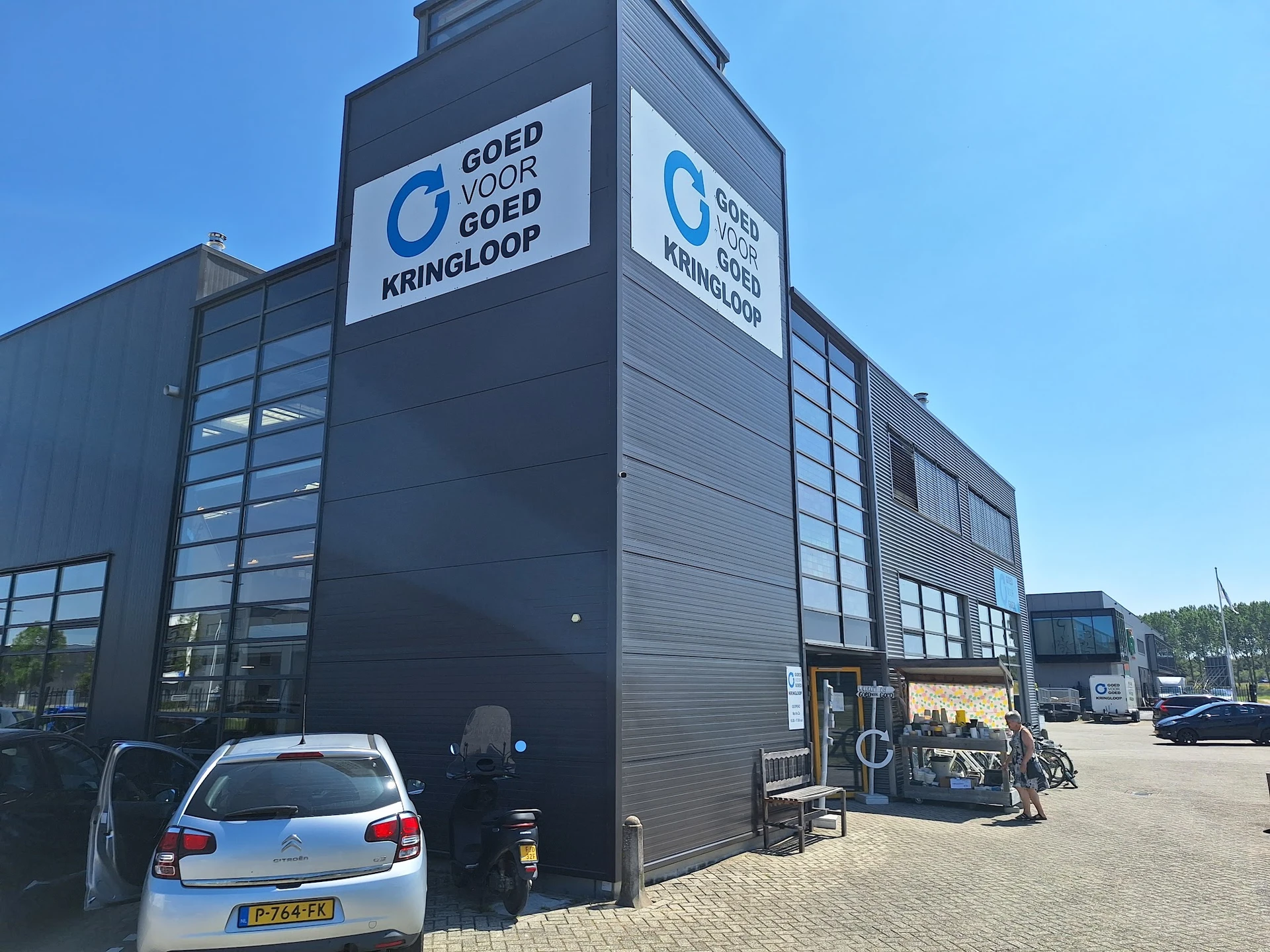 Zwart kringloopcentrum met glazen ramen en blauw logo 'Goed voor Goed Kringloop' op de voorgevel, auto geparkeerd ervoor