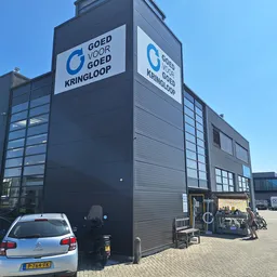 Zwart kringloopcentrum met glazen ramen en blauw logo 'Goed voor Goed Kringloop' op de voorgevel, auto geparkeerd ervoor