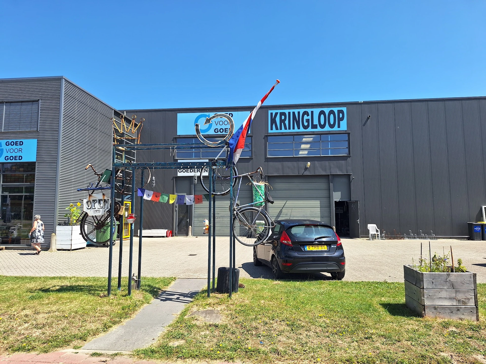 Grijze gevel van kringloopwinkel met naam KRINGLOOP op voorgevels, kunstwerk met fietsen en paal op terras ervoor
