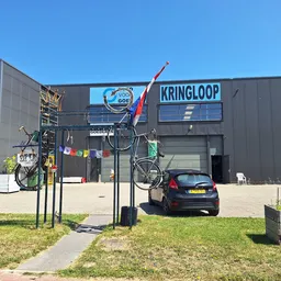 Grijze gevel van kringloopwinkel met naam KRINGLOOP op voorgevels, kunstwerk met fietsen en paal op terras ervoor