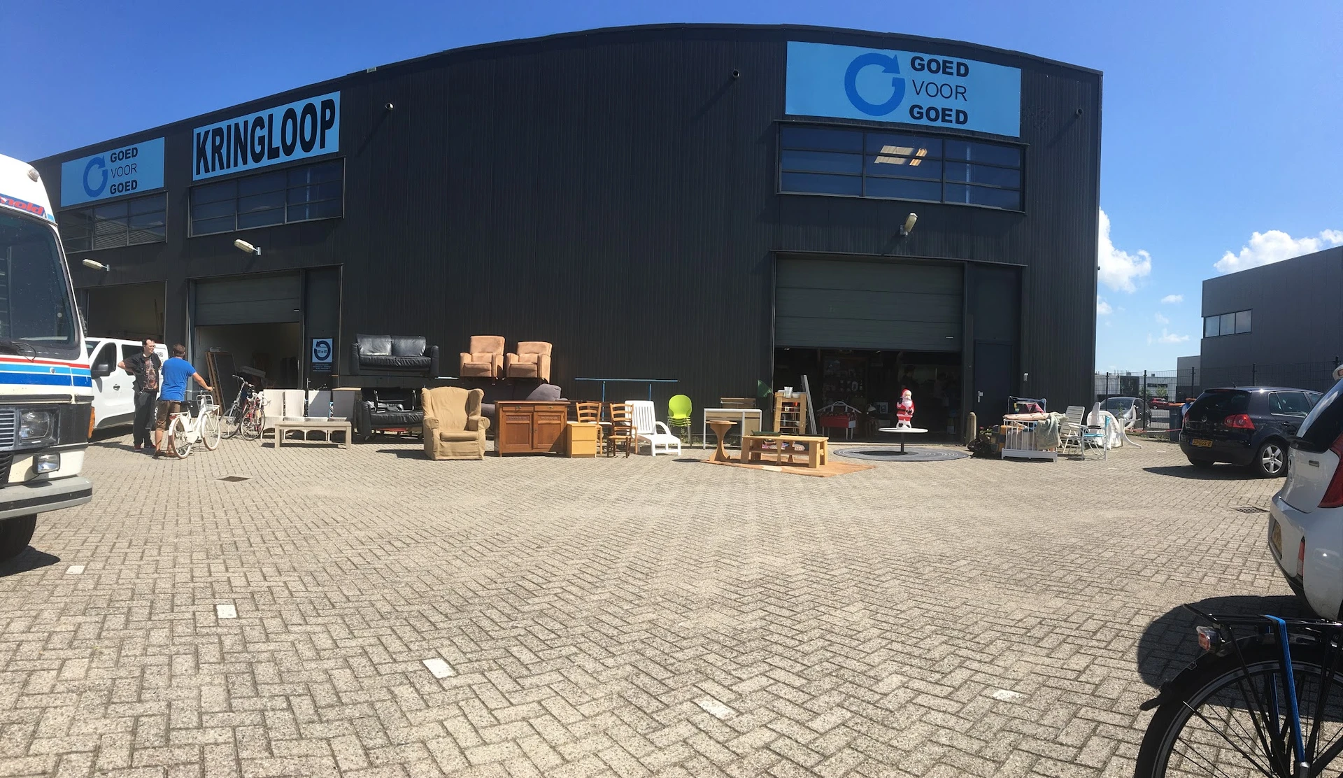 Zwart kringloopcentrum met 'KRINGLOOP' en 'GOED VOOR GOED' signage, diverse gebruikte spullen uitgestald op het plein ervoor