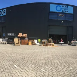Zwart kringloopcentrum met 'KRINGLOOP' en 'GOED VOOR GOED' signage, diverse gebruikte spullen uitgestald op het plein ervoor