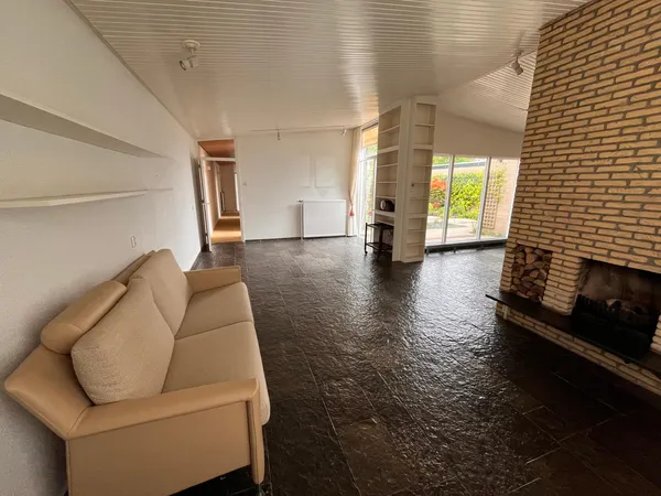 Ruim interieur van kringloopwinkel met beige bank, donkere vloer, bakstenen muur met open haard en zicht op ramen met tuin