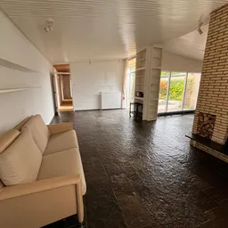 Ruim interieur van kringloopwinkel met beige bank, donkere vloer, bakstenen muur met open haard en zicht op ramen met tuin