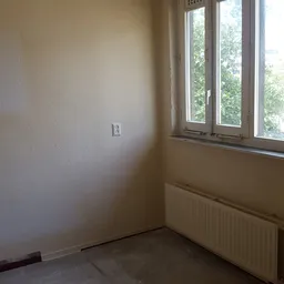 Leeg kamertje met witte wand, elektriciteitsschakelaar, radiator en raam met uitzicht op groen