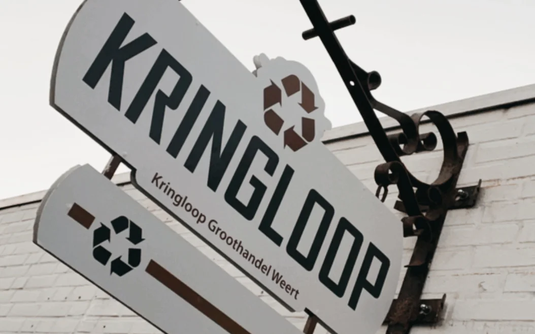 Gevelbordje van Kringloop Groothandel Weert met recyclesymbool, tegen een witgeschilderde muur met elektriciteitsleiding