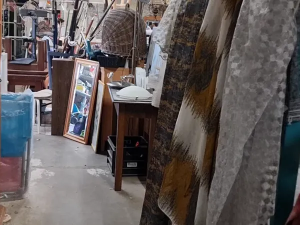 Smal gangpad tussen stellingen met diverse gebruikte spullen en meubels in een kringloopwinkel