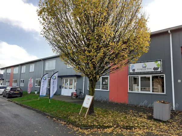 Grijze en rode gevel van kringloopwinkel met grote boom ervoor en ingang zichtbaar