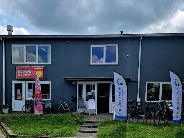 Blauwe gevel van kringloopwinkel met witte ramen, ingang, fietsen en blauwe vlaggen met logo