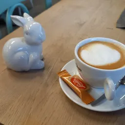Kopje koffie met latte art op schoteltje met koekje, naast een wit konijntje decoratie