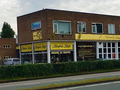 Bruine bakstenen gevel van kringloopwinkel met gele belettering en etalagevensters aan de Amsterdamsestraatweg