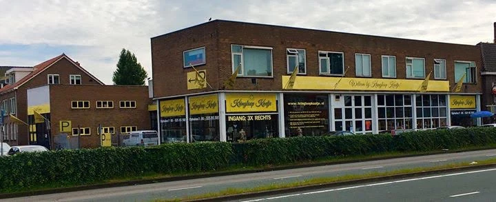 Bruine bakstenen gevel van kringloopwinkel met gele belettering en etalagevensters aan de Amsterdamsestraatweg