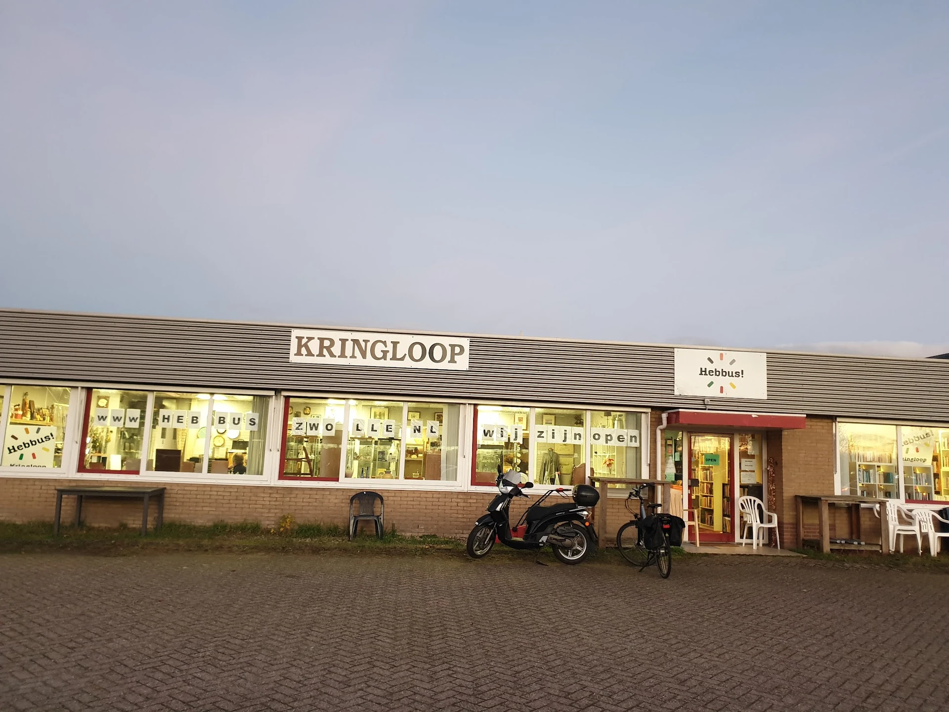 Gevel van kringloopwinkel met verlichte etalages, winkelnaam KRINGLOOP zichtbaar, scooter geparkeerd voor de ingang