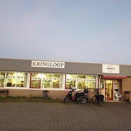 Gevel van kringloopwinkel met verlichte etalages, winkelnaam KRINGLOOP zichtbaar, scooter geparkeerd voor de ingang